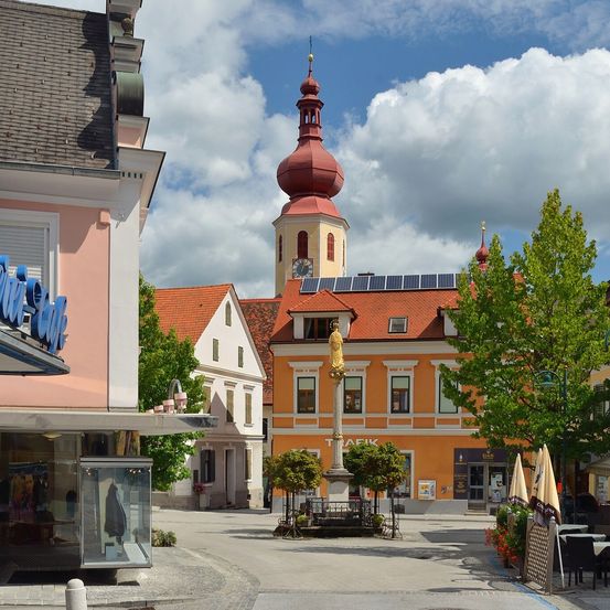 An einem sonnigen Tag zeigt ein Stadtplatz mit einer Kirche, einer Statue und Gebäuden mit Solarpanels auf ihren Dächern.