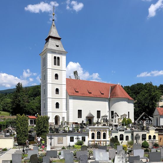 Eine große weiße Kirche mit einem roten Dach steht in einem Friedhof, umgeben von einigen kleinen weißen Gebäuden. Der Himmel ist blau mit verstreuten Wolken.