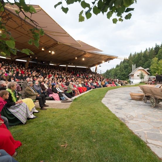 Ein Freiluft-Amphitheater, mit vielen Menschen auf Bänken unter einem Vordach. Ein Rasenbereich mit einem Steinweg führt zu einem kleinen Haus. Ein Karren ist rechts geparkt.
