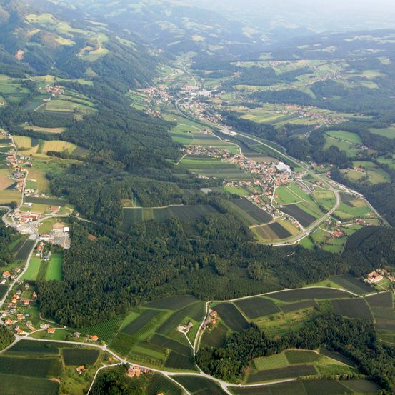 Luftaufnahme eines Tals mit einer Mischung aus grünen Feldern, Wohngebieten und einer kurvenreichen Straße. Die Landschaft umfasst üppige Wälder und kleine Städte, die zwischen den Hügeln verstreut sind.
