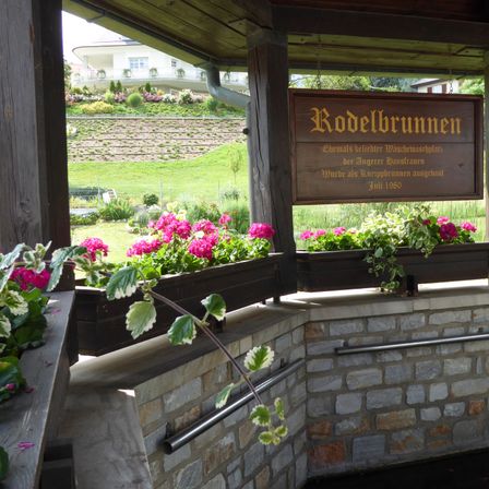 Ein hölzernes Schild namens Rodelbrunnen hängt über zwei Holzpflanzgefäßen mit rosa Blumen. Das Schild sagt, dass es im Juli 1980 als Brunnen gebaut wurde.