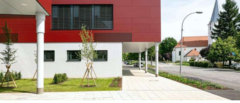 Ein modernes rotes Gebäude mit Glasfenstern und einem Gehweg davor. Im Vordergrund befinden sich Bäume, Büsche und eine Straßenlampe.
