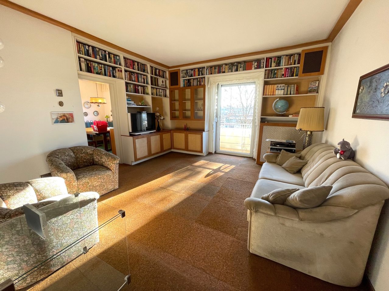 Ein sonniges Wohnzimmer mit Bücherregalen, einem Fernseher und einem Glastisch. Es gibt eine Couch, einen Sessel und eine Lampe auf der rechten Seite.