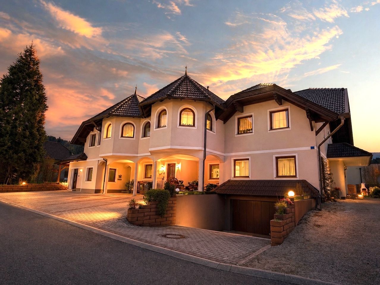 A large house with several windows and doors lit up at dusk. The house has a garage and plants in front.
