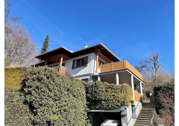 Ein zweistöckiges Haus mit Balkon und Stufen, die zum Eingang führen, umgeben von Büschen und Bäumen unter einem klaren blauen Himmel.