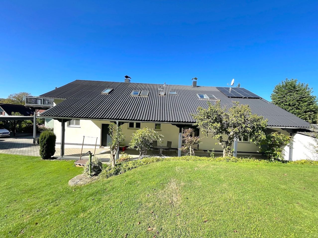 Ein modernes Haus mit einem Schieferdach und Solarmodulen. Es hat eine Veranda mit einem Tisch und Stühlen. Das Haus ist von einem gepflegten Rasen und mehreren Bäumen umgeben.