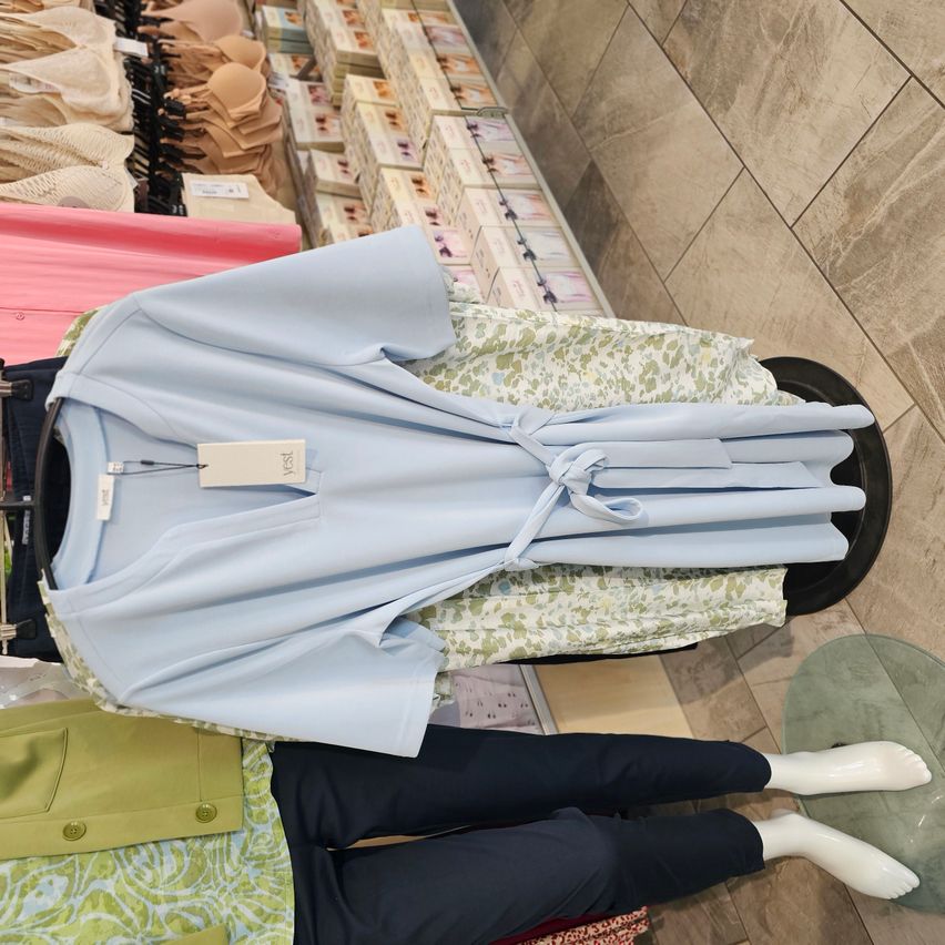 Ein hellblaues Oberteil mit Bindegürtel, gefaltet und auf einem Mannequin in einem Bekleidungsgeschäft mit gefliestem Boden und verschiedenen Kleidungsstücken im Hintergrund ausgestellt.