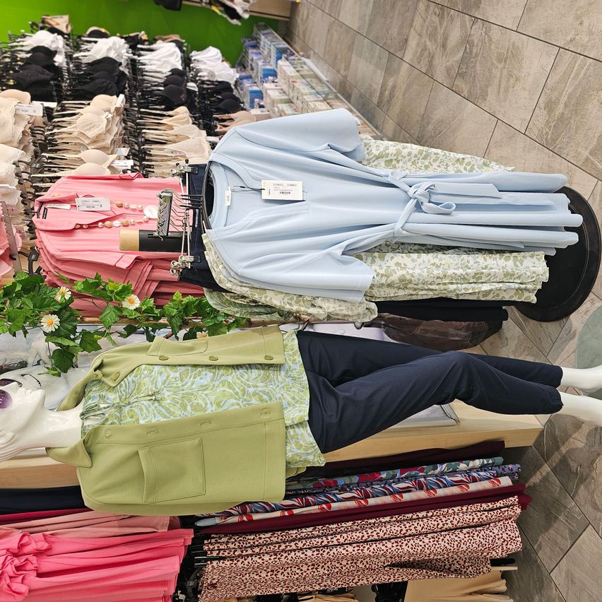 Ein Bekleidungsgeschäft mit Mannequins, die verschiedene Outfits präsentieren. Ein Mannequin mit einer grünen Jacke und blauen Hosen ist prominent. Der Laden ist gefüllt mit ordentlich organisierten Kleidungsstücken und Accessoires an Kleiderbügeln.