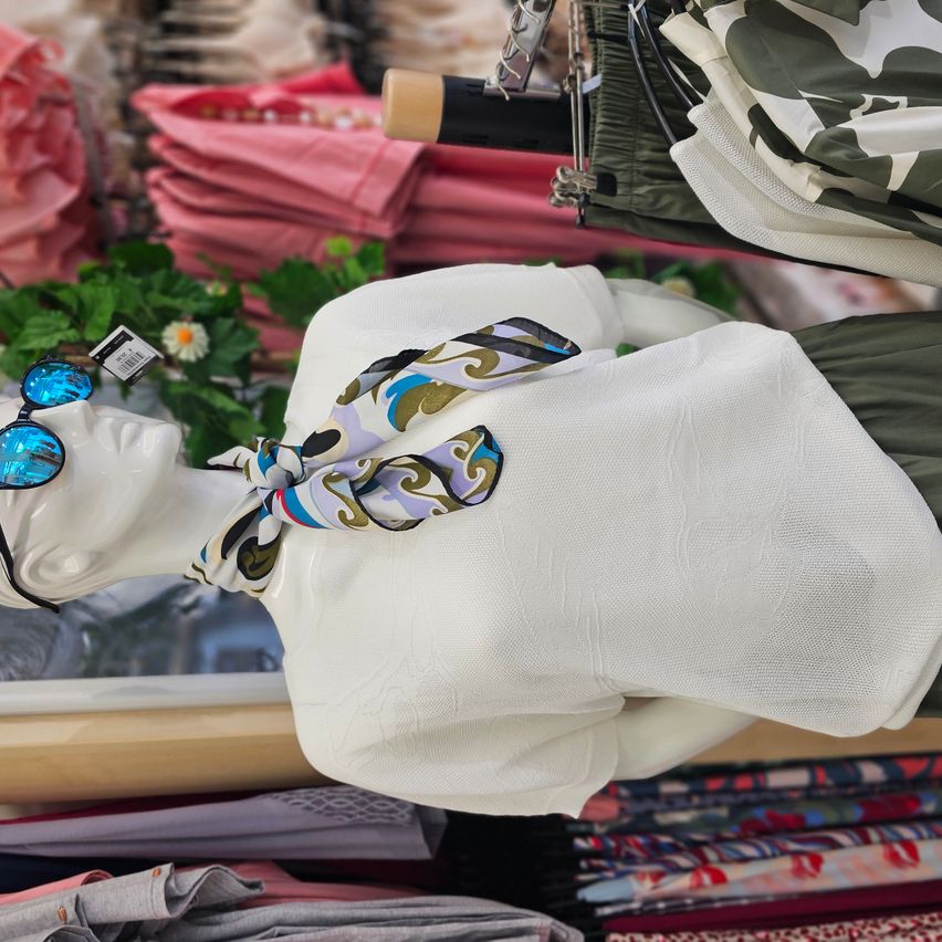 Eine Puppe in einem weißen Oberteil mit einem gemusterten Schal wird in einem Bekleidungsgeschäft ausgestellt. Dahinter hängen verschiedene Hemden an Kleiderbügeln, und ein grüner Tarnstoff ist sichtbar.