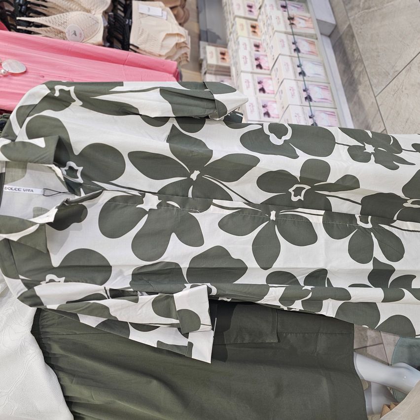 Eine grüne Bluse mit Blumenmuster von Dolce Vita wird auf einem Mannequin präsentiert. Die Bluse liegt über einem grünen Rock und einem weißen Kissen. Dahinter sind Regale mit rosa und weißen Schachteln zu sehen.