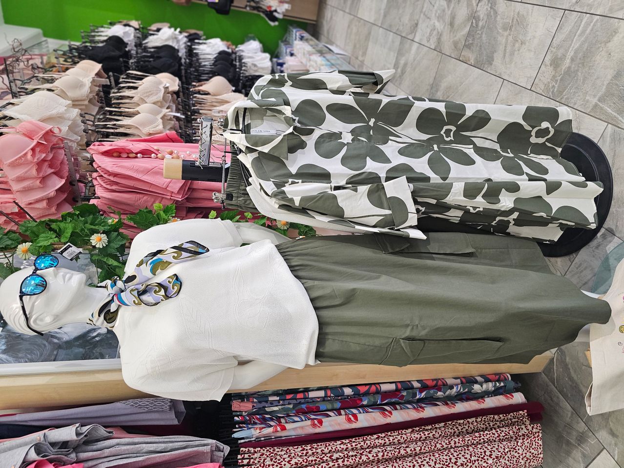 Ein Bekleidungsgeschäft mit einer Bluse mit Blumendruck auf einem Mannequin, einer weißen Bluse und grünen Hosen, zusammen mit Stapeln von rosa und schwarzer Unterwäsche an Kleiderbügeln.