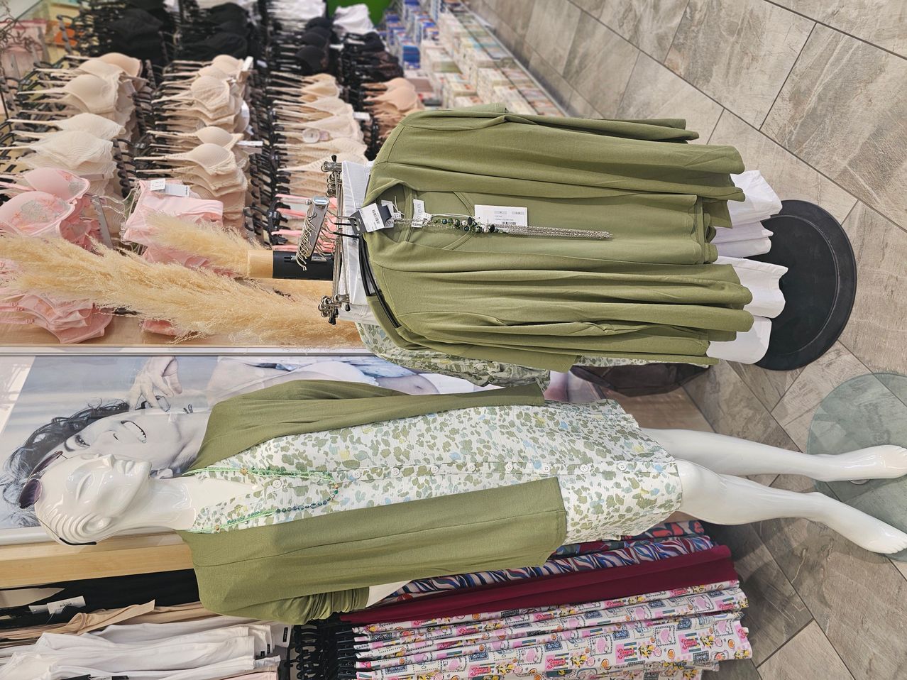 Ein Bekleidungsgeschäft zeigt eine Puppe in einem grünen Blumenkleid und einer Jacke, mit Regalen voller Kleidung im Hintergrund.