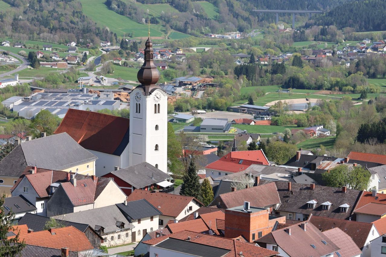 Eine Luftaufnahme einer Stadt mit einem Kirchturm und einer Uhr, umgeben von verschiedenen Gebäuden, Bäumen und Grünflächen, mit einer Brücke in der Ferne.