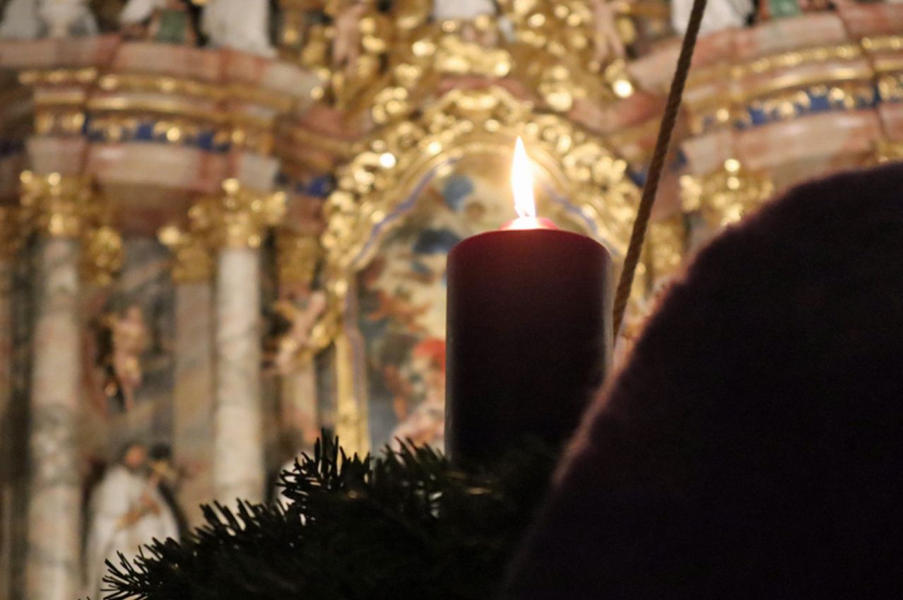 Eine brennende Kerze steht auf einem geschmückten Weihnachtsbaum in einer Kirche, mit einem goldenen Altar und Kunstwerken im Hintergrund.