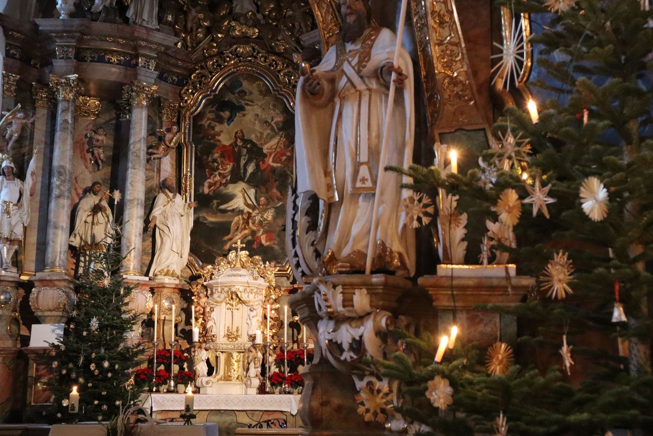 Ein kunstvoller Altaraufsatz in einer Kirche zeigt ein Gemälde von Engeln und Heiligen. Eine Statue eines Bischofs steht in der Nähe und hält einen Stab. Kerzen schmücken den Altar, und ein Weihnachtsbaum mit Dekorationen ist im Vordergrund.