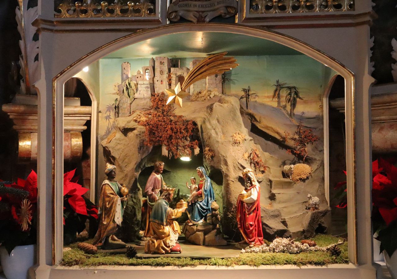 Eine Krippenszene mit Figuren von Maria, Josef und dem Jesuskind in einer Höhle, umgeben von Felsen und Grünpflanzen, mit einem goldenen Stern darüber und einer Kulisse einer Stadt.