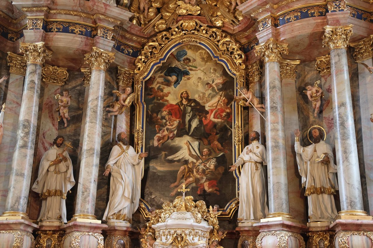 Ein großes religiöses Gemälde mit mehreren Figuren ist in einer Kirche ausgestellt. Es ist mit Gold- und Blauverzierungen gerahmt und wird von zwei Statuen flankiert. Die Statuen sind ebenfalls in weißen Gewändern gekleidet.