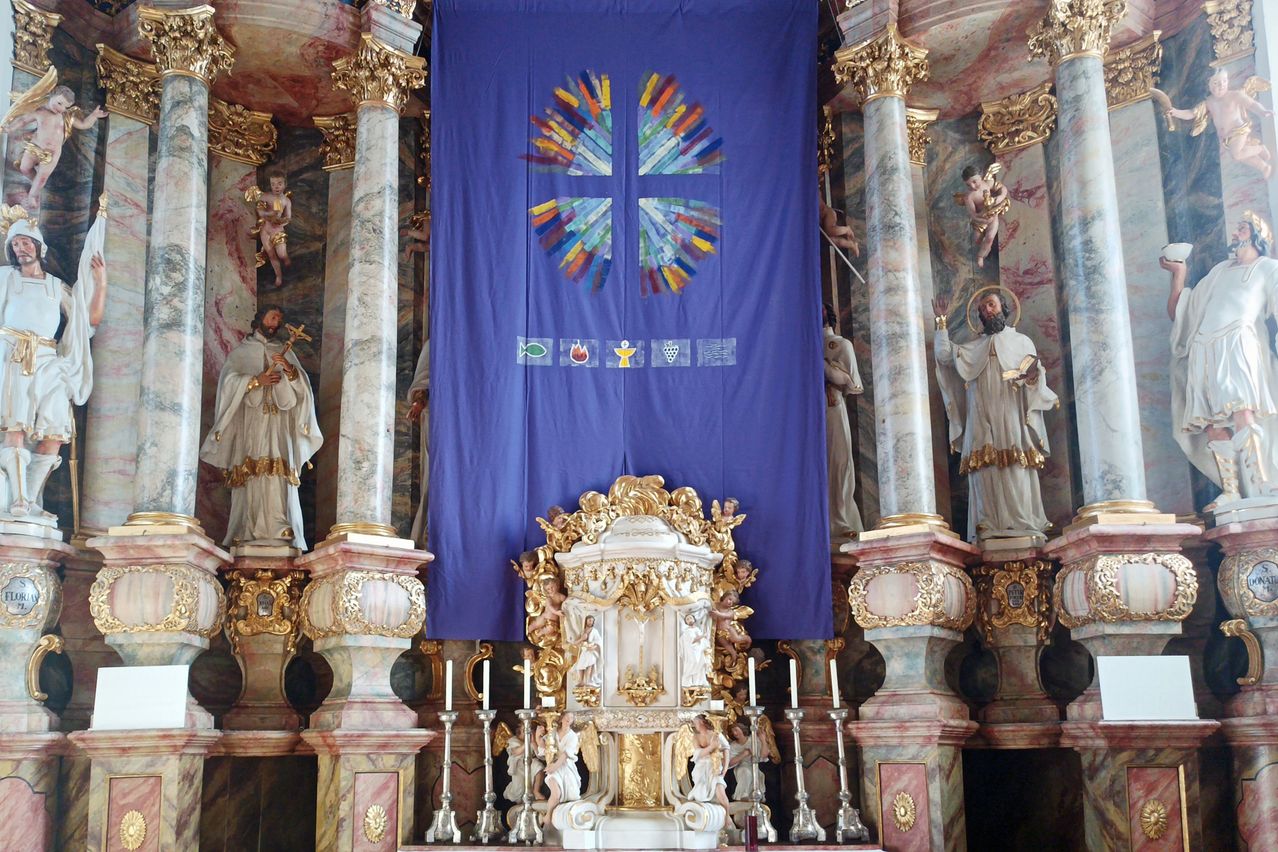 Ein großes blaues Banner mit einem Kreuz und religiösen Symbolen hängt über einem golden verzierten Altar in einer Kirche. Kerzen flankieren den Altar, und Statuen von Heiligen säumen die Wände.