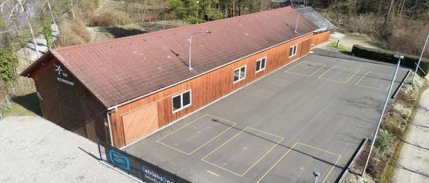 Luftaufnahme eines Gebäudes mit einem braunen Dach und mehreren Fenstern. Das Gebäude ist von einem leeren Parkplatz mit freien Parkplätzen umgeben. Im Hintergrund sind Bäume und eine Straße zu sehen.
