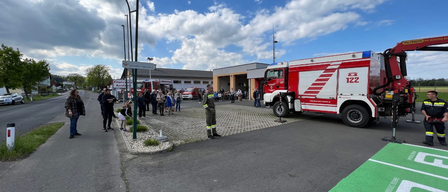 Ein Feuerwehrauto steht auf der Straße geparkt, während eine Menge Menschen zusieht. Die Straße hat einen Bordstein und Straßenlaternen. Bäume und Gebäude sind im Hintergrund.