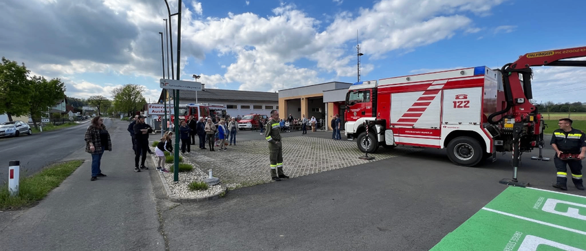 Ein Feuerwehrauto steht auf der Straße geparkt, während eine Menge Menschen zusieht. Die Straße hat einen Bordstein und Straßenlaternen. Bäume und Gebäude sind im Hintergrund.