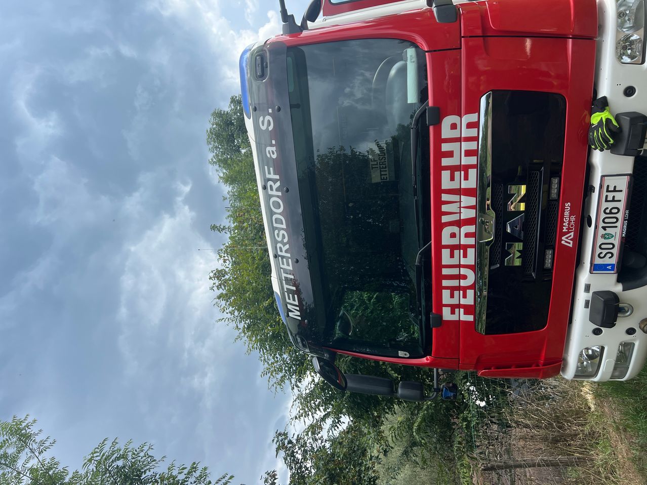 Ein Feuerwehrfahrzeug ist unter einem bewölkten Himmel geparkt. An der Seite des Fahrzeugs steht 'Feuerwehr Mettersdorf a. S.' und vorne ist die Nummer '42' zu sehen.
