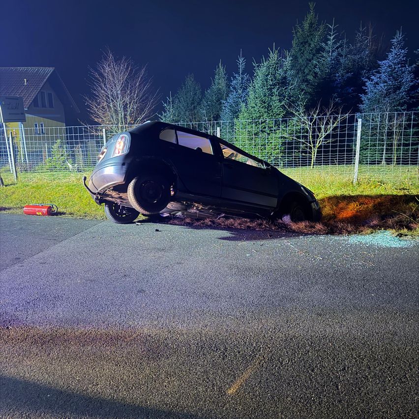 Ein schwarzes Auto ist am Straßenrand demoliert, mit einem Feuerlöscher in der Nähe. Es ist Nacht, und das Auto befindet sich in der Nähe eines Zauns mit einem Haus und Bäumen dahinter.