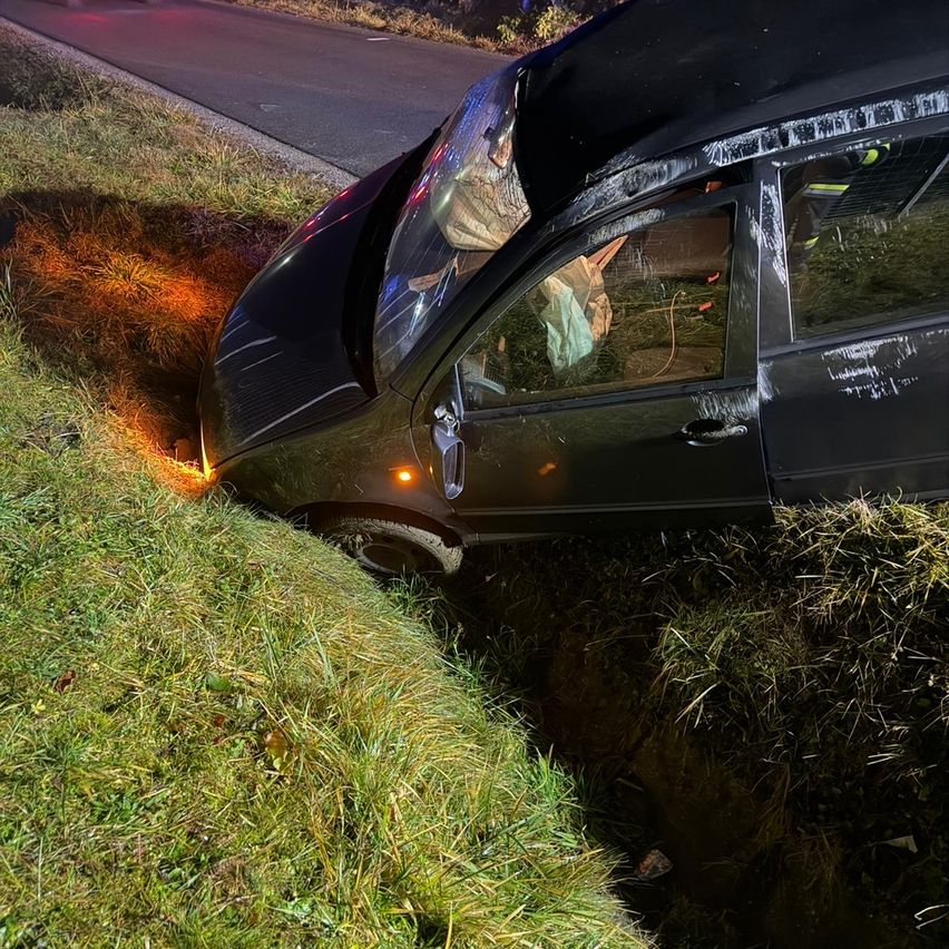 Ein schwarzes Auto ist auf der Straßenseite umgekippt. Die Front ist über einem Graben geneigt, die Autotür ist offen und die Motorhaube ist zerdrückt.