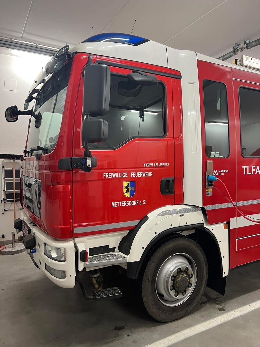 Ein roter Feuerwehrwagen mit weißem Streifen steht in einer Garage. Es steht Freiwillige Feuerwehr und TGM15.290 auf der Seite.
