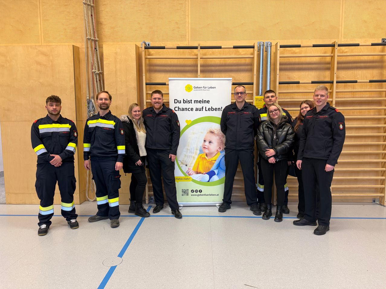 Eine Gruppe von Menschen in Uniformen steht in einem Raum, lächelt und posiert für ein Foto mit einem Banner in der Mitte, das ein Kind und den Text 'Du bist meine Chance auf Leben!' zeigt. Das Banner ist blau und grün.