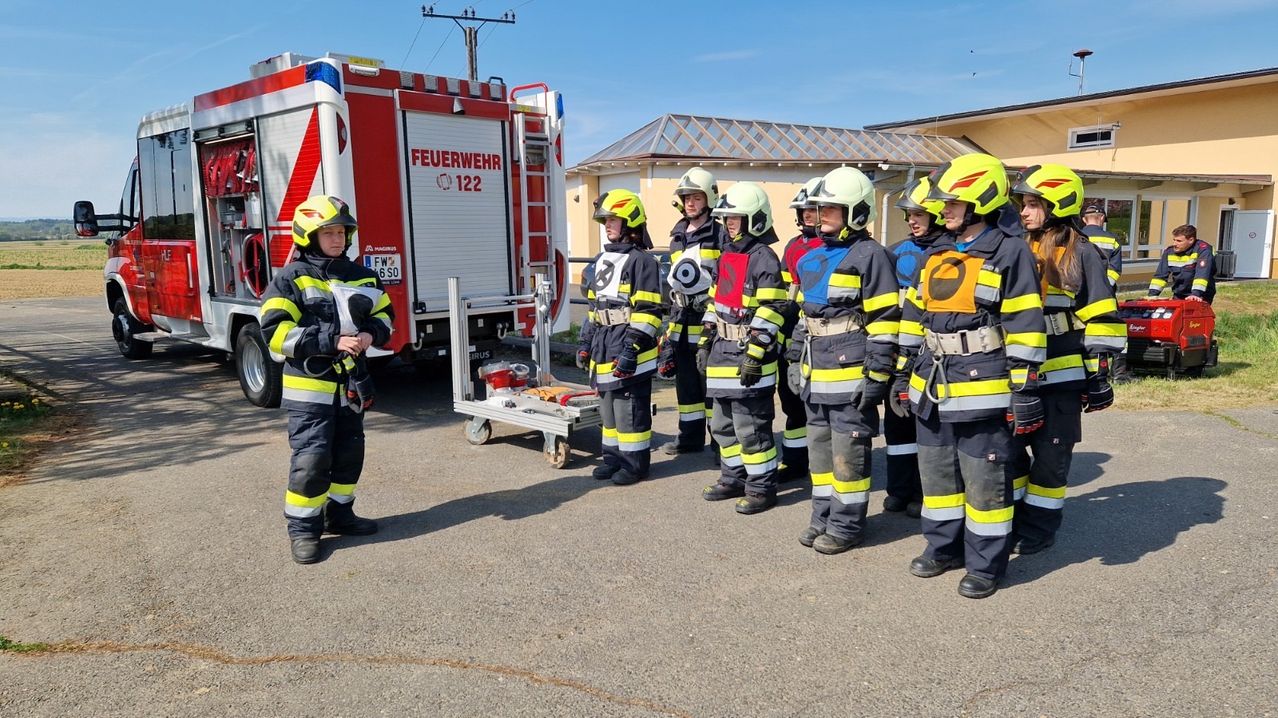 A group of firefighters in uniform stands in a line outside near a fire truck.
