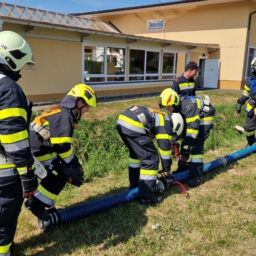 Firefighters in full gear are working on a large blue hose on a grassy field, with a building in the background.