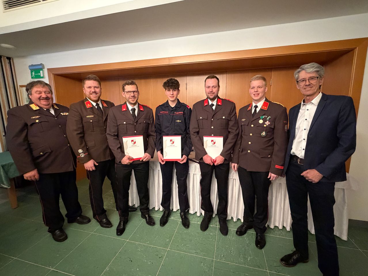 Seven individuals in military uniforms, standing together, hold certificates. They smile, likely at an award ceremony. A white tablecloth is behind them.