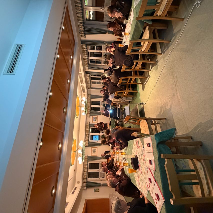 An aerial view of a dining hall with people sitting at long tables with green tablecloths, engaged in conversation. The room has wooden walls, glass windows, and mounted lights.