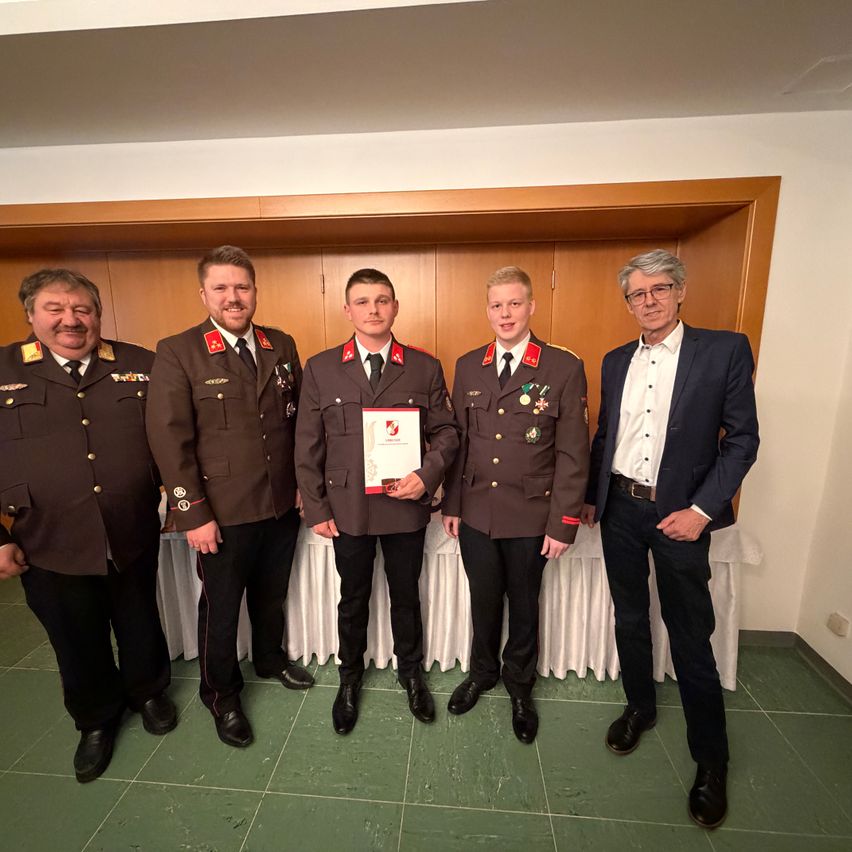 Five men in military uniforms stand in front of a white backdrop, smiling. Two hold medals, and a man in a suit stands on the right.