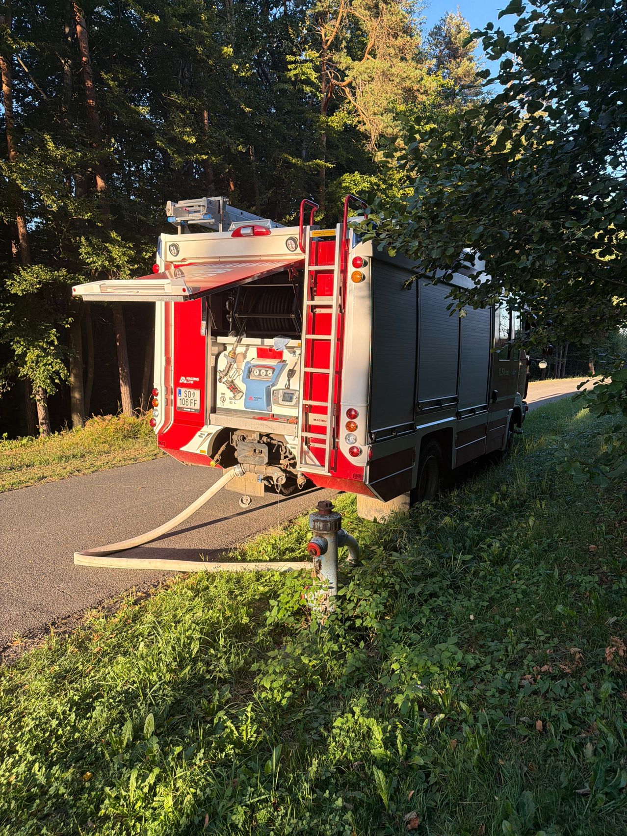A fire truck is parked on the side of a road, with its rear door open and a hose connected to a fire hydrant. The truck has a license plate reading 'SO 4 106 FF'.