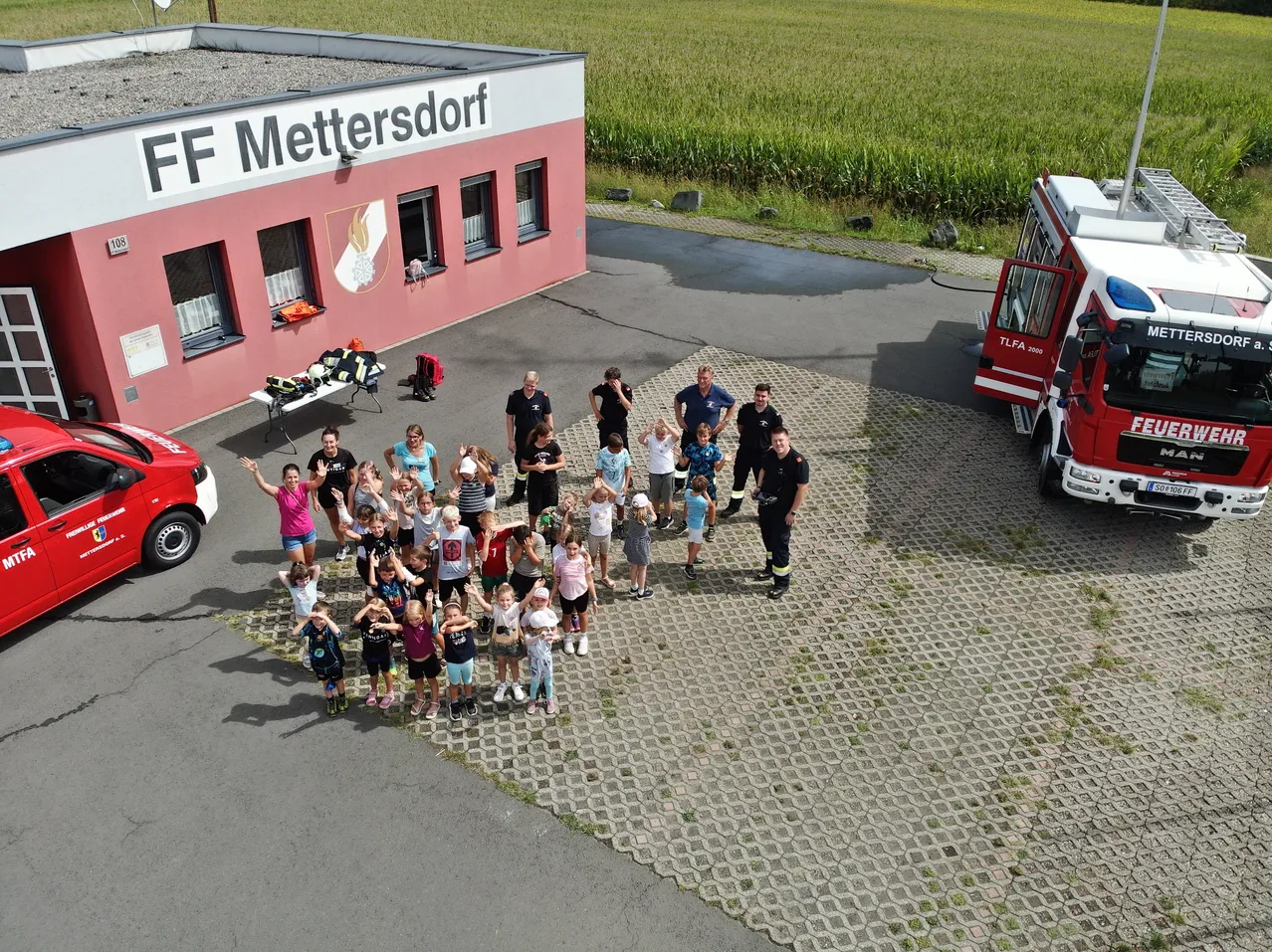 Eine Gruppe von Kindern und Erwachsenen versammelt sich vor der Feuerwehrstation FF Mettersdorf, wobei Feuerwehrleute und ein Feuerwehrauto im Hintergrund zu sehen sind.