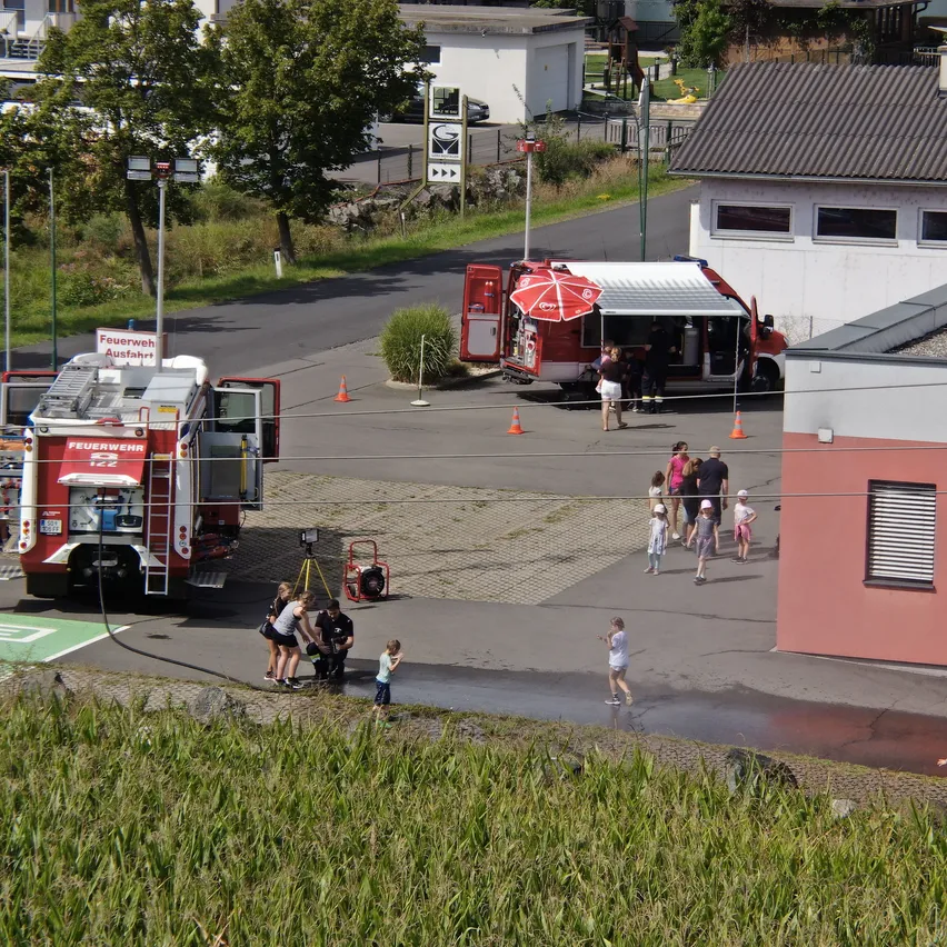 Ein Feuerwehrauto ist am Straßenrand geparkt, mit ausgefahrener Leiter. Ein roter LKW mit offenem Verdeck ist in der Nähe geparkt. Mehrere Menschen stehen herum, einige in der Nähe des Feuerwehrwagens.