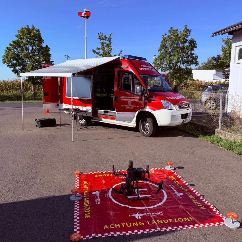 Ein roter Feuerwehrwagen mit offener Tür ist am Straßenrand geparkt. Ein Drohnenmodell liegt auf einem roten Landezonen-Matte. Ein Vordach ist neben dem Wagen aufgestellt. Bäume und ein Gebäude sind im Hintergrund.
