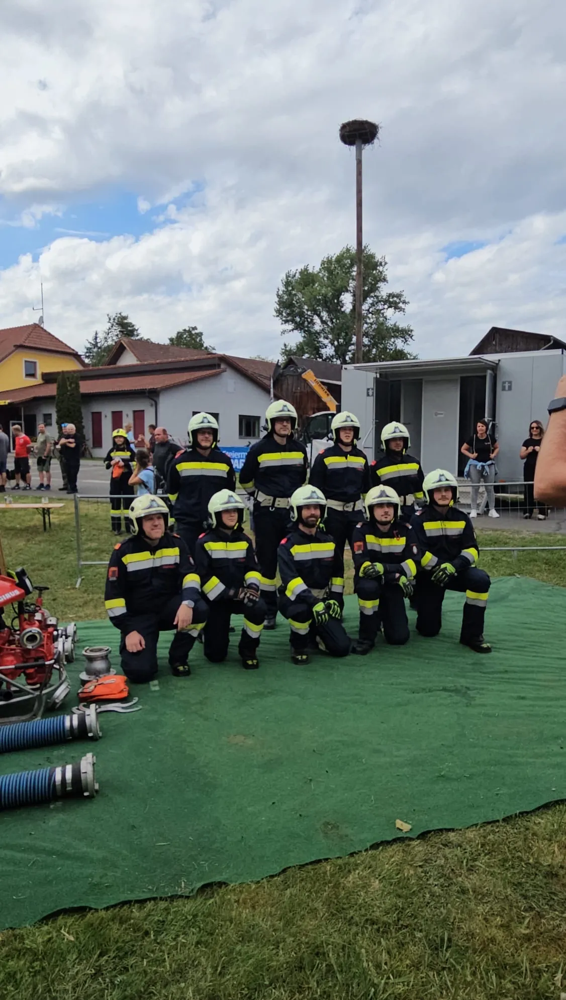 Eine Gruppe Feuerwehrleute in Uniform posiert für ein Foto auf einem grünen Feld mit Feuerwehrausrüstung im Hintergrund.