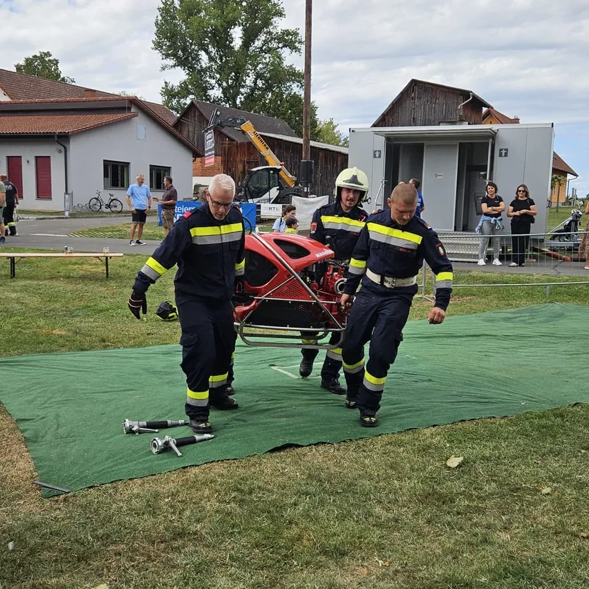 Drei Feuerwehrleute tragen eine Trage mit einem roten Motorrad darauf. Sie tragen Uniformen und Helme. Hinter einem Zaun stehen Menschen und beobachten.