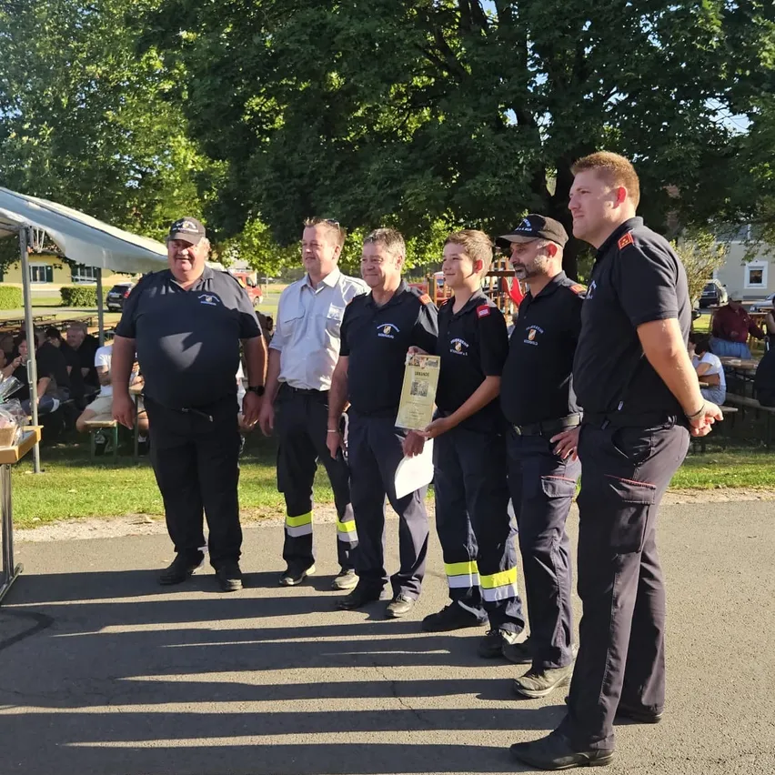 Fünf Feuerwehrleute stehen in einer Reihe, einer hält ein Zertifikat, mit einer Menge und Zelten im Hintergrund.