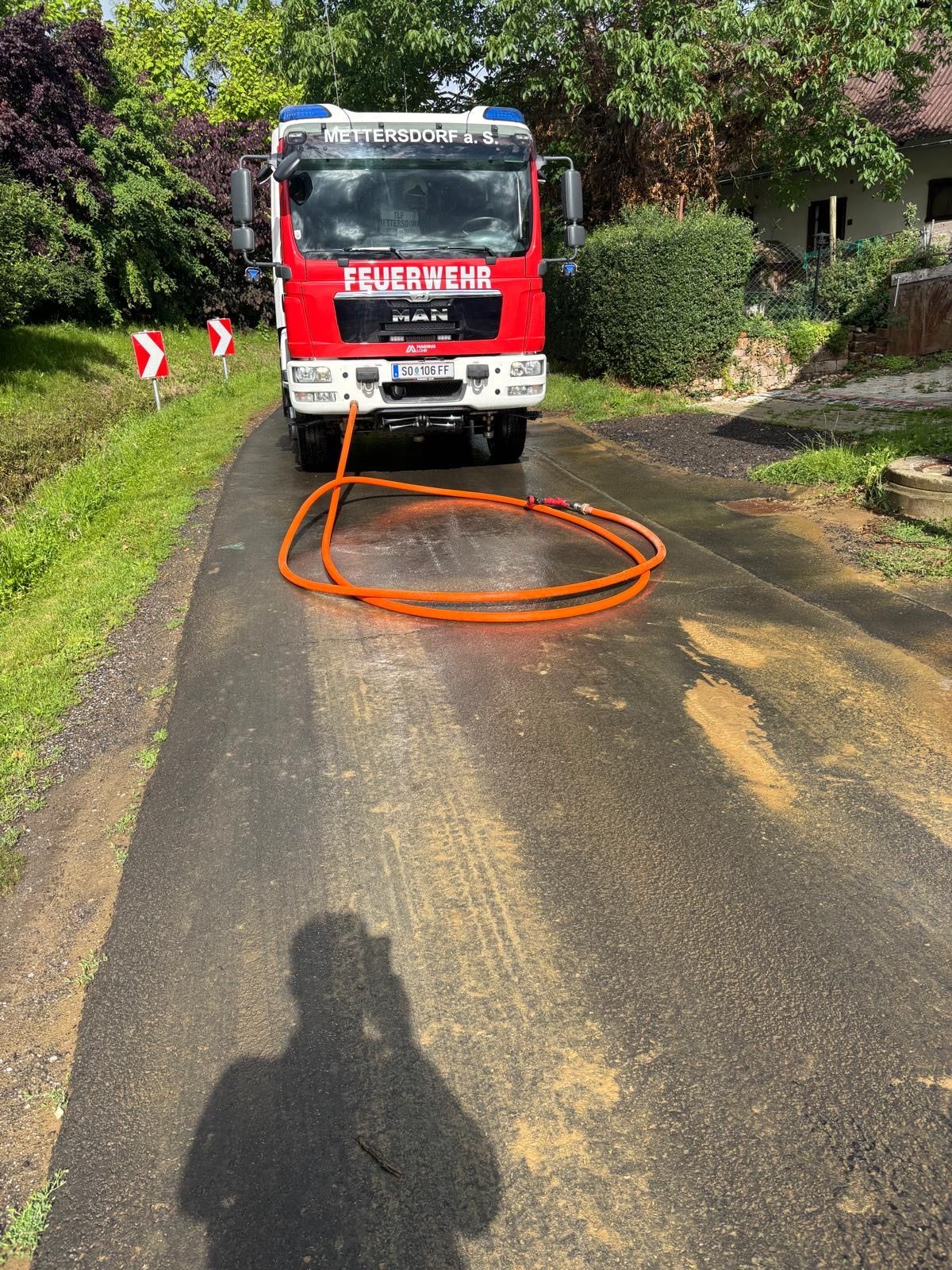 A red fire truck is parked on a wet road with a hose on the side, near bushes and a house.