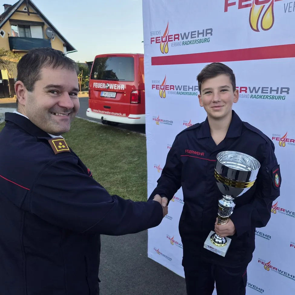 Ein Feuerwehrmann und ein junger Junge in Uniform schütteln sich die Hände vor einem Banner mit dem Text 'Feuerwehr'. Der Junge hält einen Pokal. Dahinter steht ein roter Van und ein Haus.