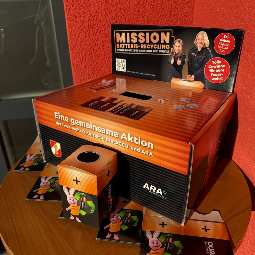 Eine Pappschachtel mit der Aufschrift 'Mission Batterie-Recycling'. Die Schachtel ist orange und schwarz und hat ein Loch in der Mitte. Es gibt auch einige Karten mit einem Hasencharakter.