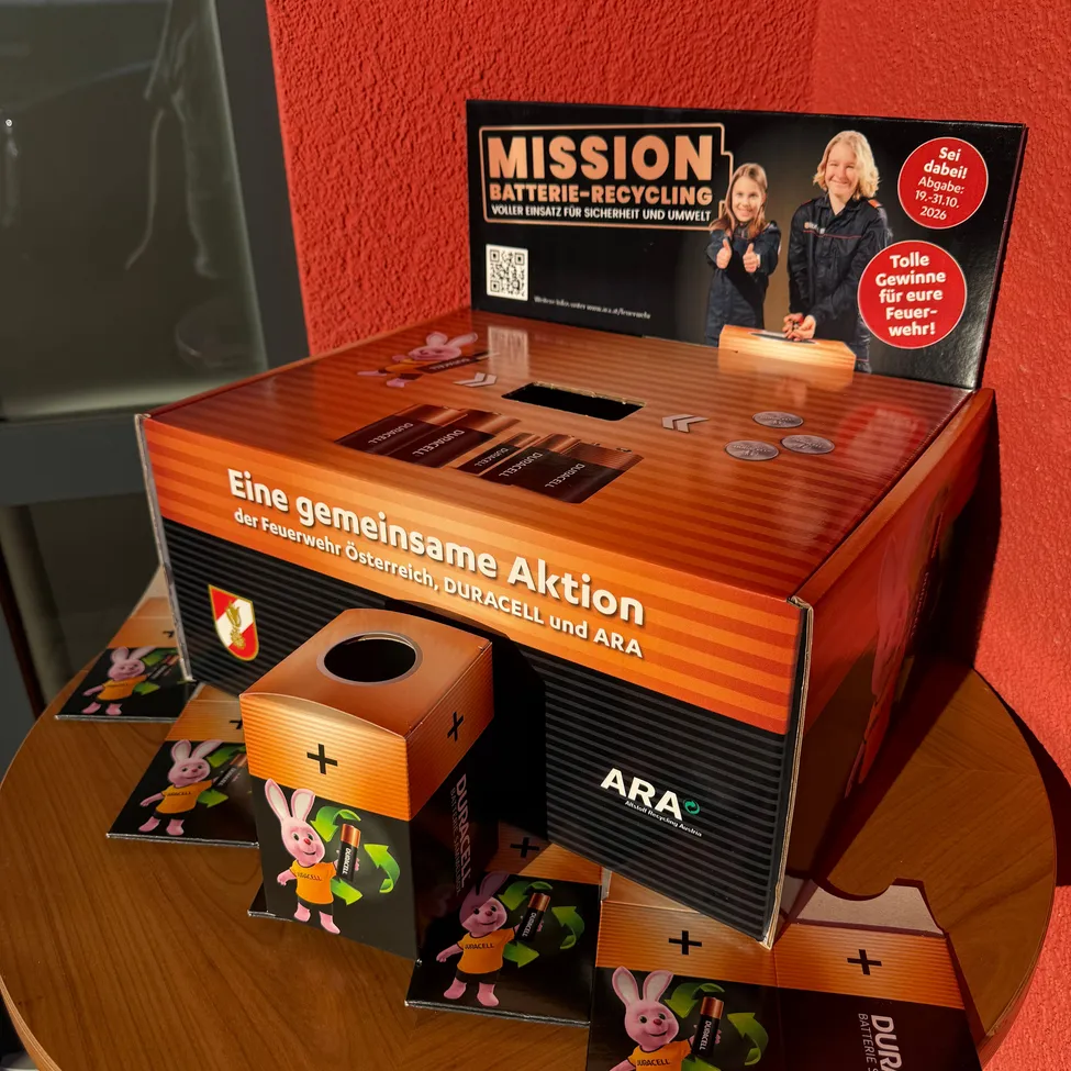 Eine Pappschachtel mit der Aufschrift 'Mission Batterie-Recycling'. Die Schachtel ist orange und schwarz und hat ein Loch in der Mitte. Es gibt auch einige Karten mit einem Hasencharakter.