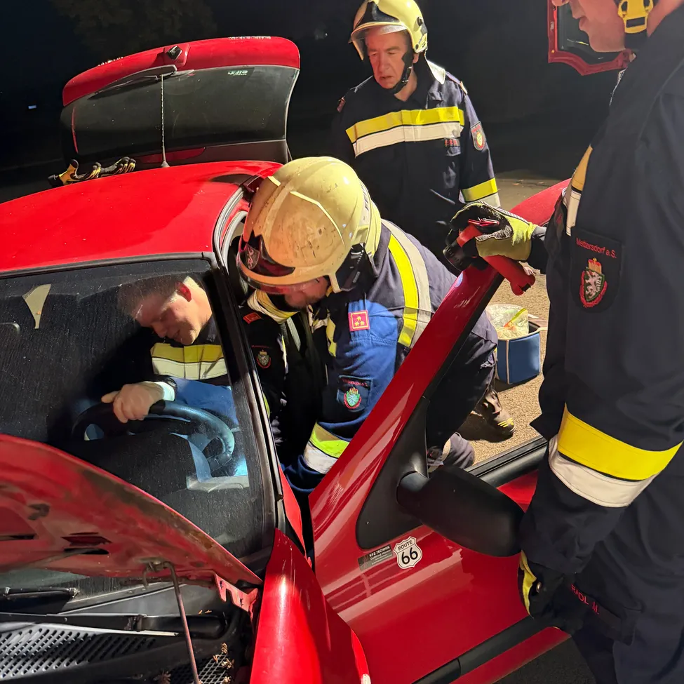 Drei Feuerwehrleute arbeiten nachts an einem roten Auto. Einer sitzt auf dem Fahrersitz, ein anderer arbeitet am Motor und der dritte hält ein Werkzeug.