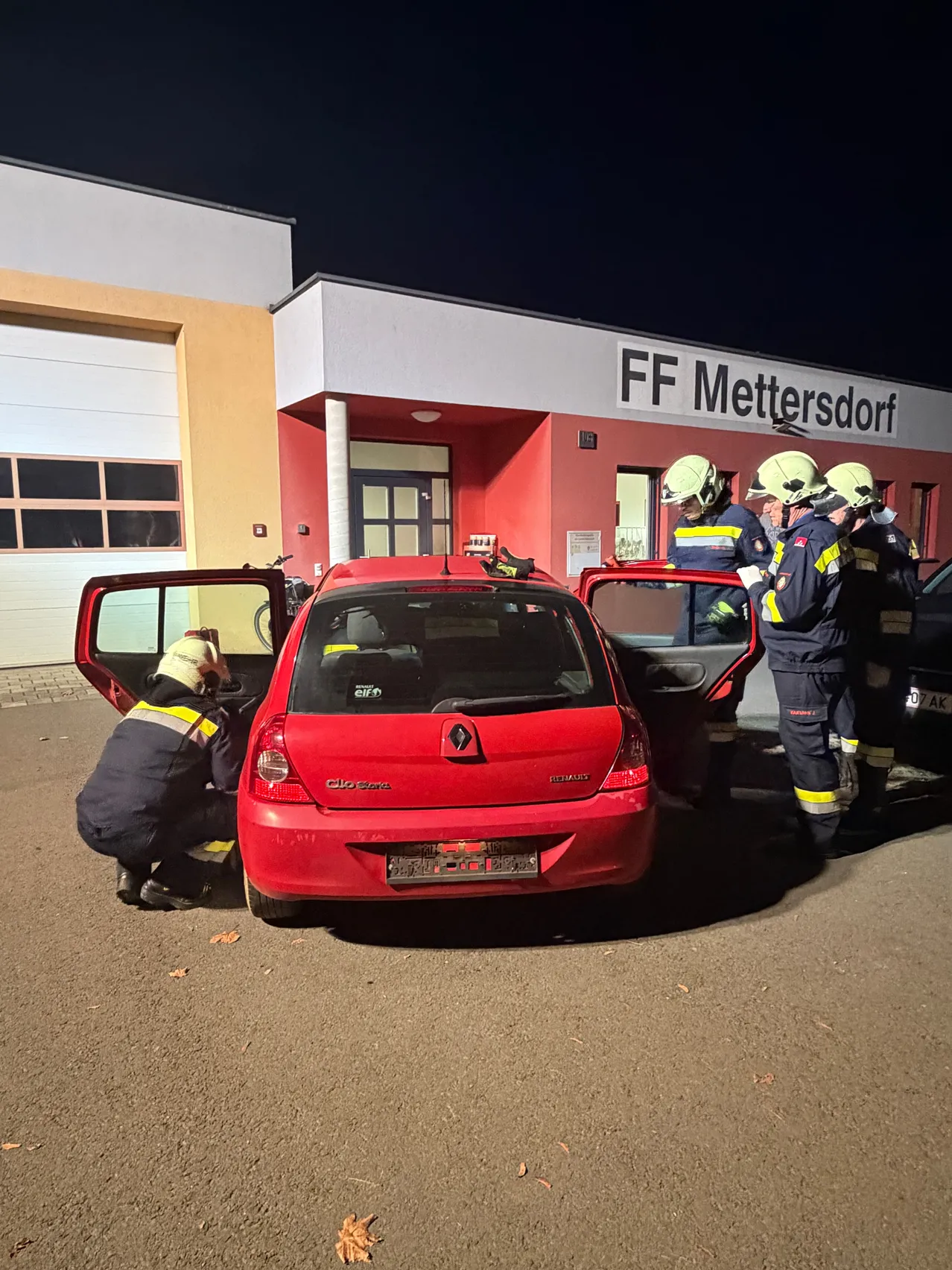 Eine Gruppe von Feuerwehrleuten arbeitet an einem roten Renault Clio Auto vor einem Feuerwehrhaus in der Nacht.