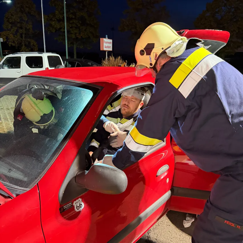 Zwei Feuerwehrleute kümmern sich um einen Mann in einem roten Auto. Einer trägt einen gelben Helm und der andere einen blauen Helm. Der Mann sitzt auf dem Fahrersitz mit einem Verband auf dem Kopf.
