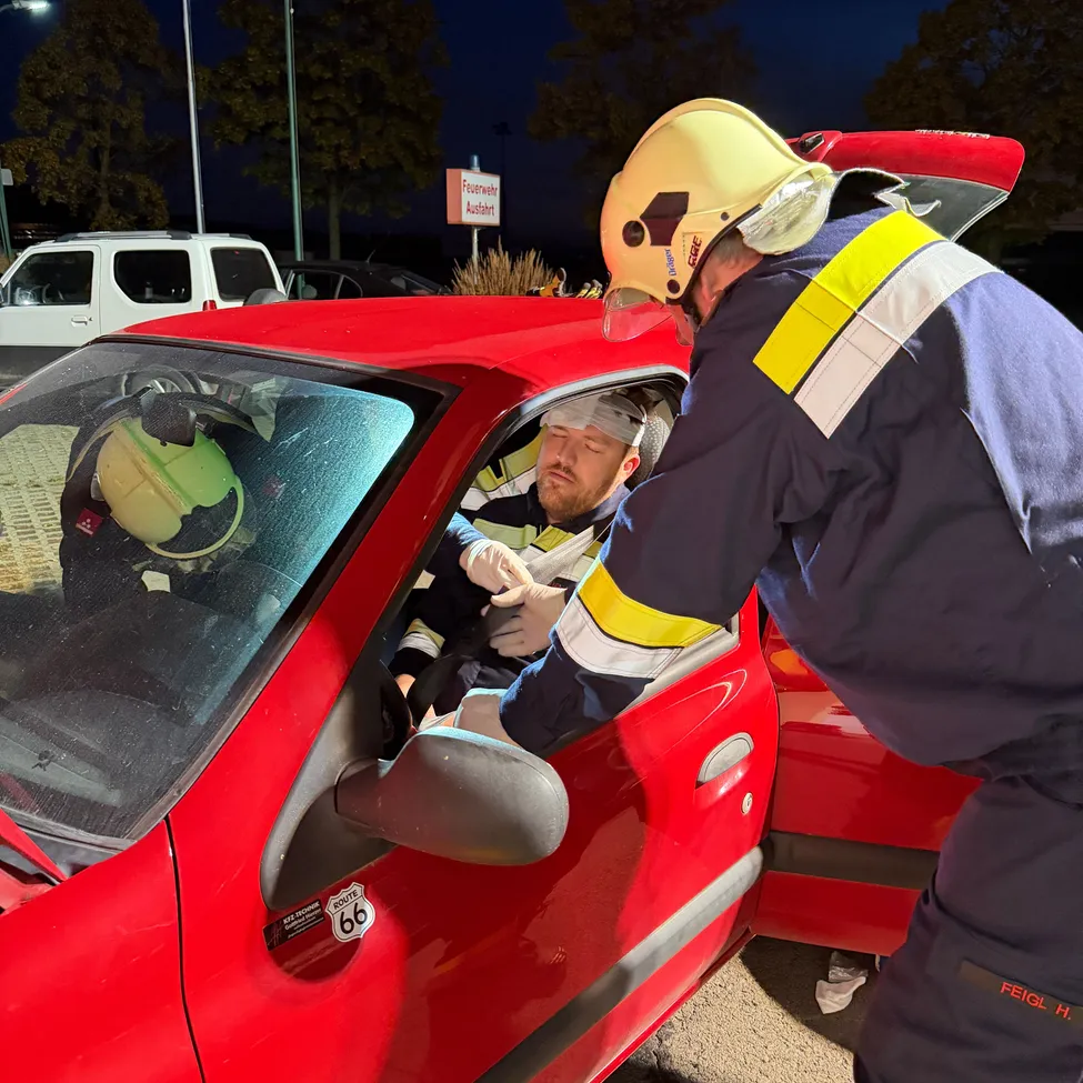 Zwei Feuerwehrleute kümmern sich um einen Mann in einem roten Auto. Einer trägt einen gelben Helm und der andere einen blauen Helm. Der Mann sitzt auf dem Fahrersitz mit einem Verband auf dem Kopf.